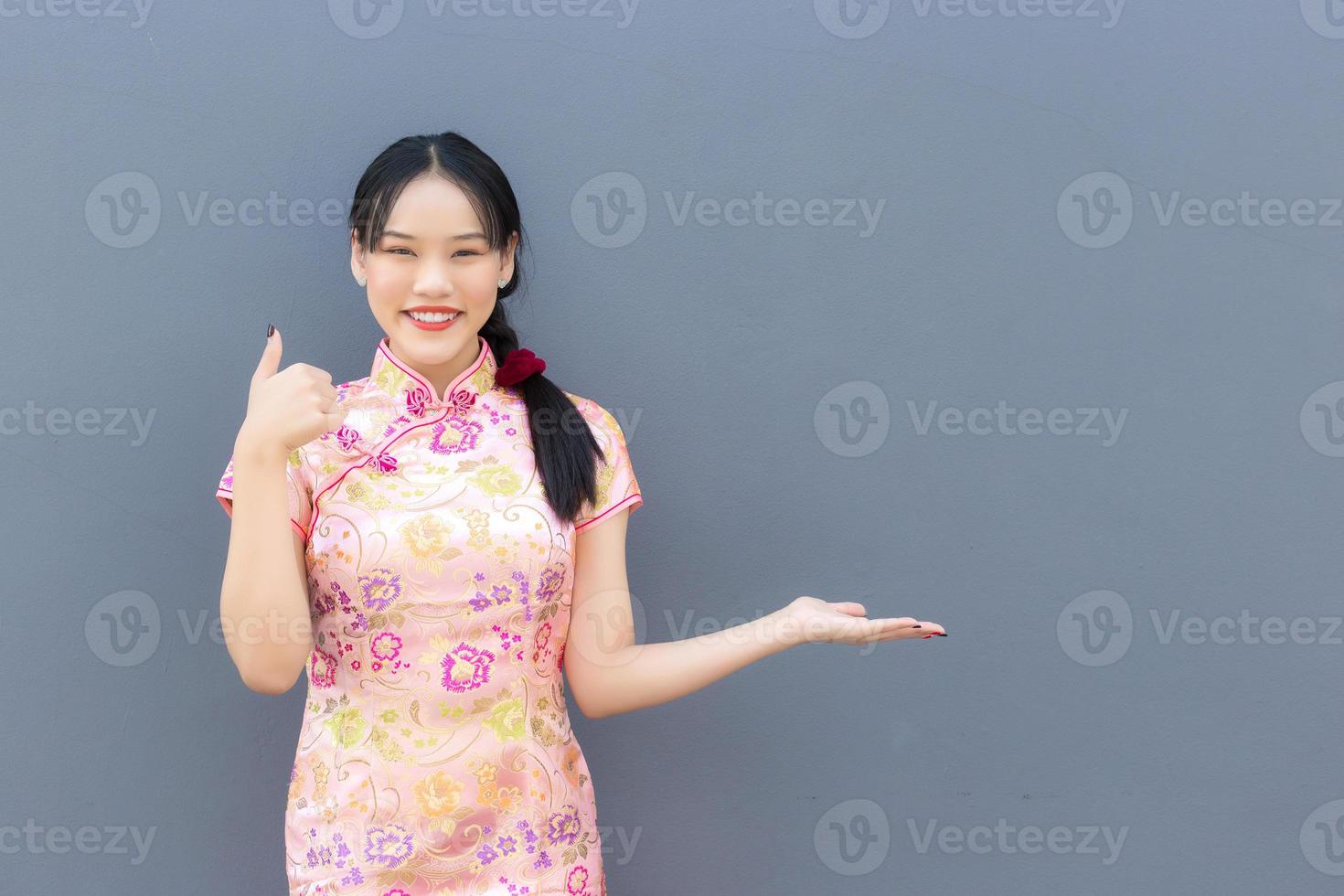 Beautiful Asian woman with long hair who wears pink Cheongsam dress in Chinese new year theme while her hand shows to present something and showing hand as thump up and looks at camera. photo