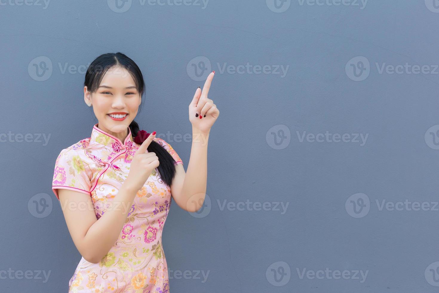 hermosa mujer asiática con cabello largo que usa un vestido rosa cheongsam en el tema del año nuevo chino mientras su mano muestra algo y muestra la mano para señalar y mira a la cámara sonriendo. foto