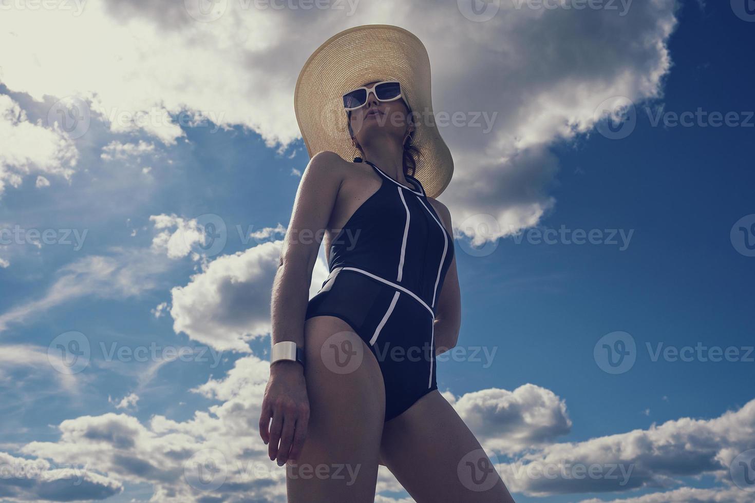 ortrait of a woman in a swimsuit, hat and sunglasses in summer on the riverbank against the sky photo