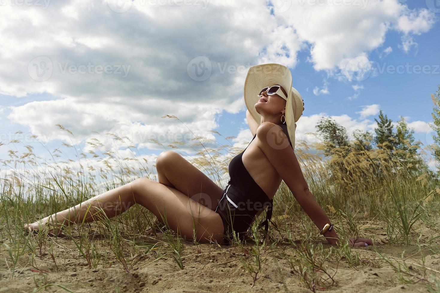woman in a swimsuit, hat and sunglasses sunbathes in summer on the riverbank among the grass photo