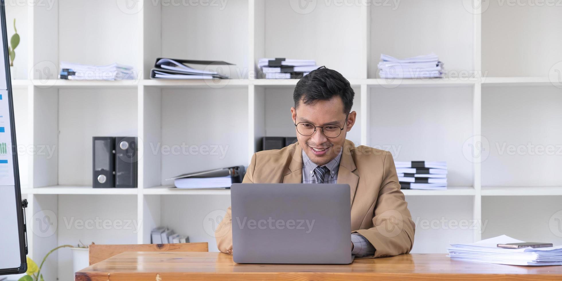 Happy young businessman in suit looking at laptop excited by good news online, lucky successful winner man standing at office desk raising hand in yes gesture celebrating business success win result photo