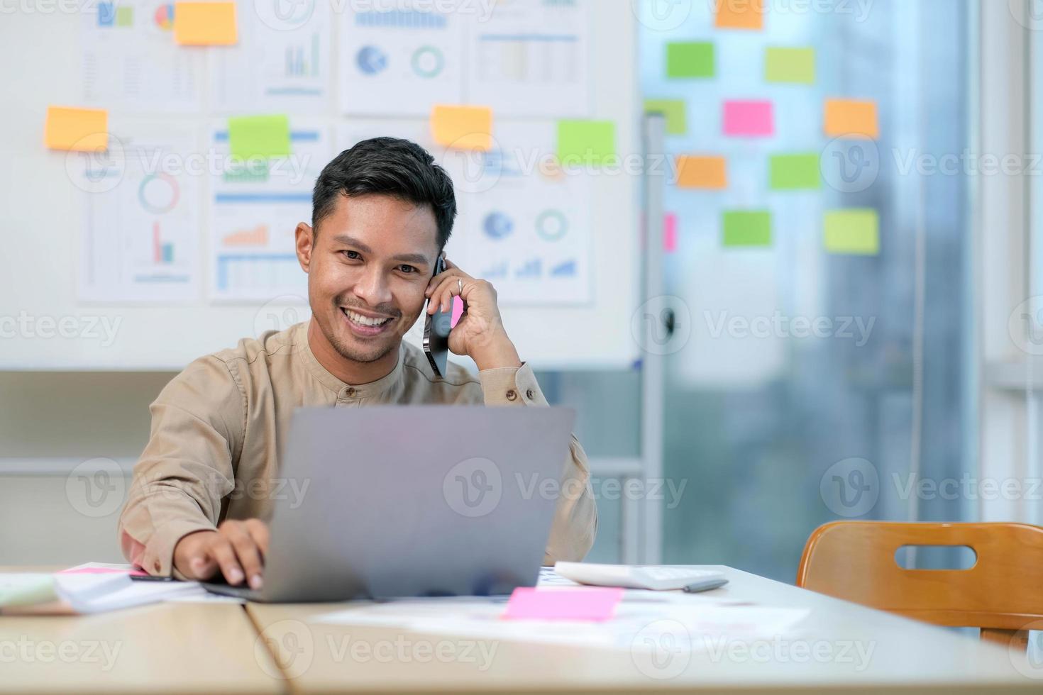 apuesto joven empleado sentado en el escritorio y mirando el monitor de la computadora mientras responde una llamada en la oficina foto