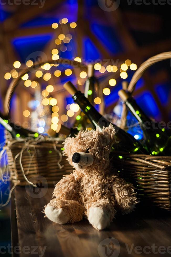 Teddy bear, champagne bottle with Christmas basket, gold garlands on dark brown wooden background, new year celebration concept photo