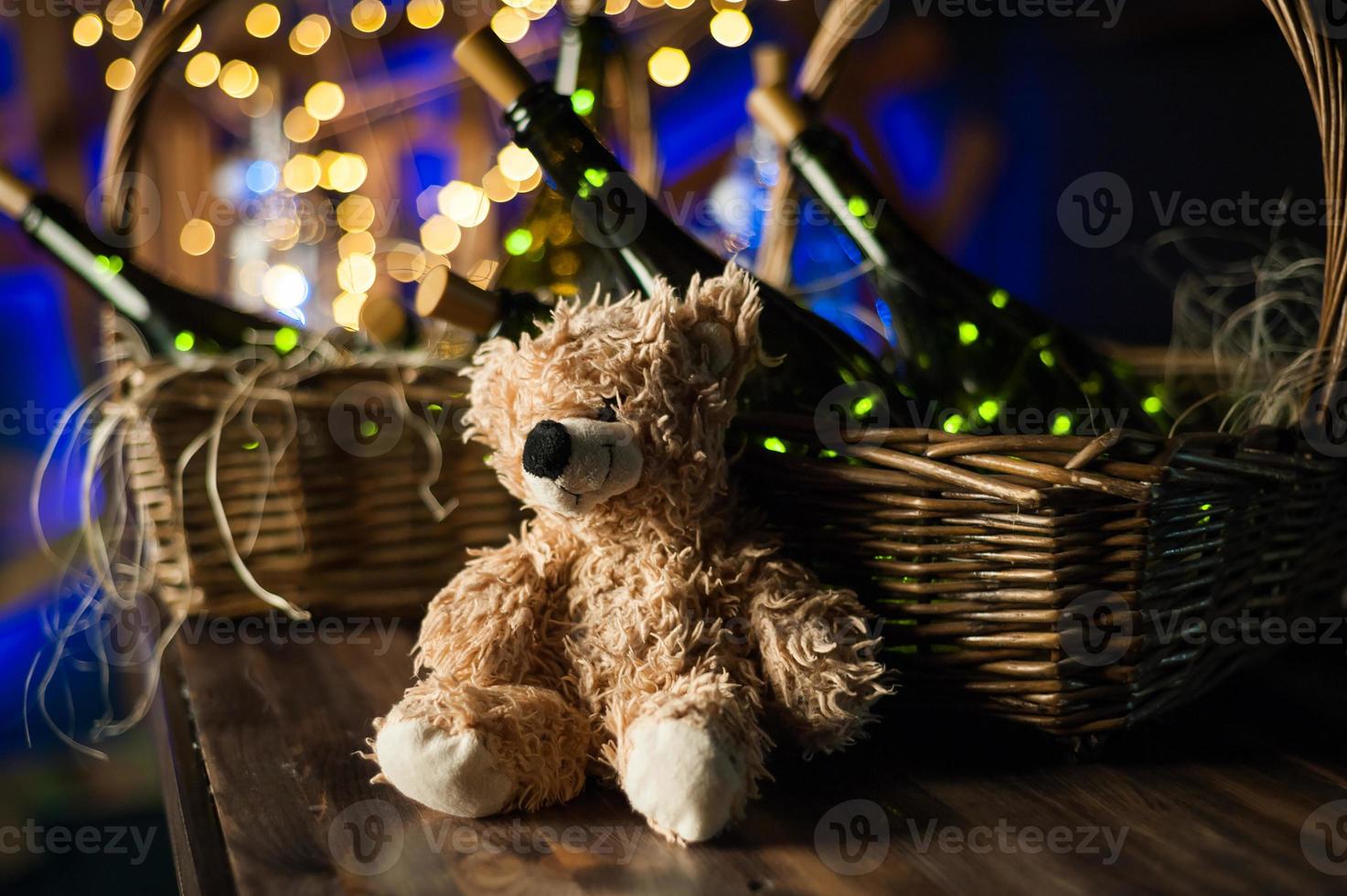 Teddy bear, champagne bottle with Christmas basket, gold garlands on dark brown wooden background, new year celebration concept photo