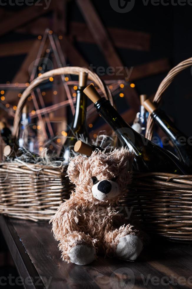 Teddy bear, champagne bottle with Christmas basket, gold garlands on dark brown wooden background, new year celebration concept photo