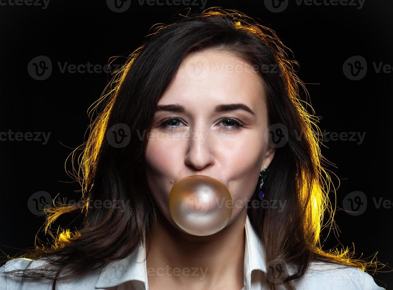 A beautiful dark-haired girl inflates a bubble of yellow gum on a black background photo