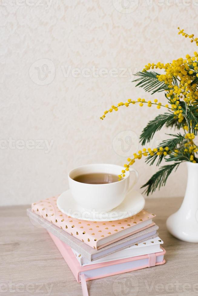 Bouquet of mimosa in a white vase, a cup of tea on a stack of notebooks for notes photo