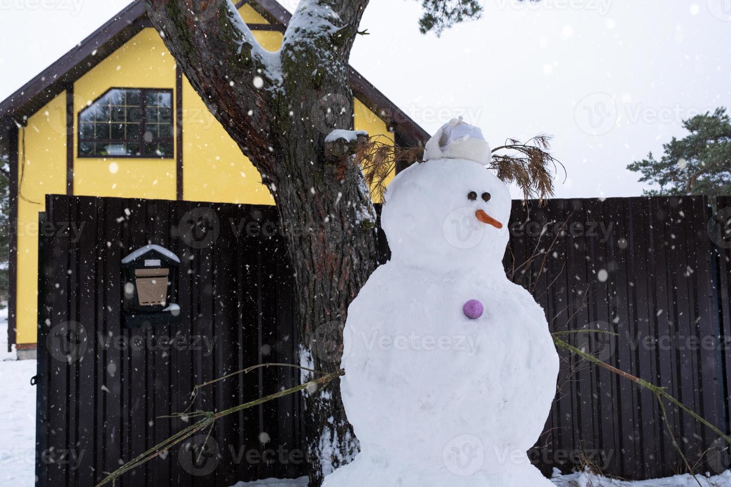 Funny snowman in a hat and scarf on the background of a yellow house in the yard. Winter, winter entertainment, snowfall photo