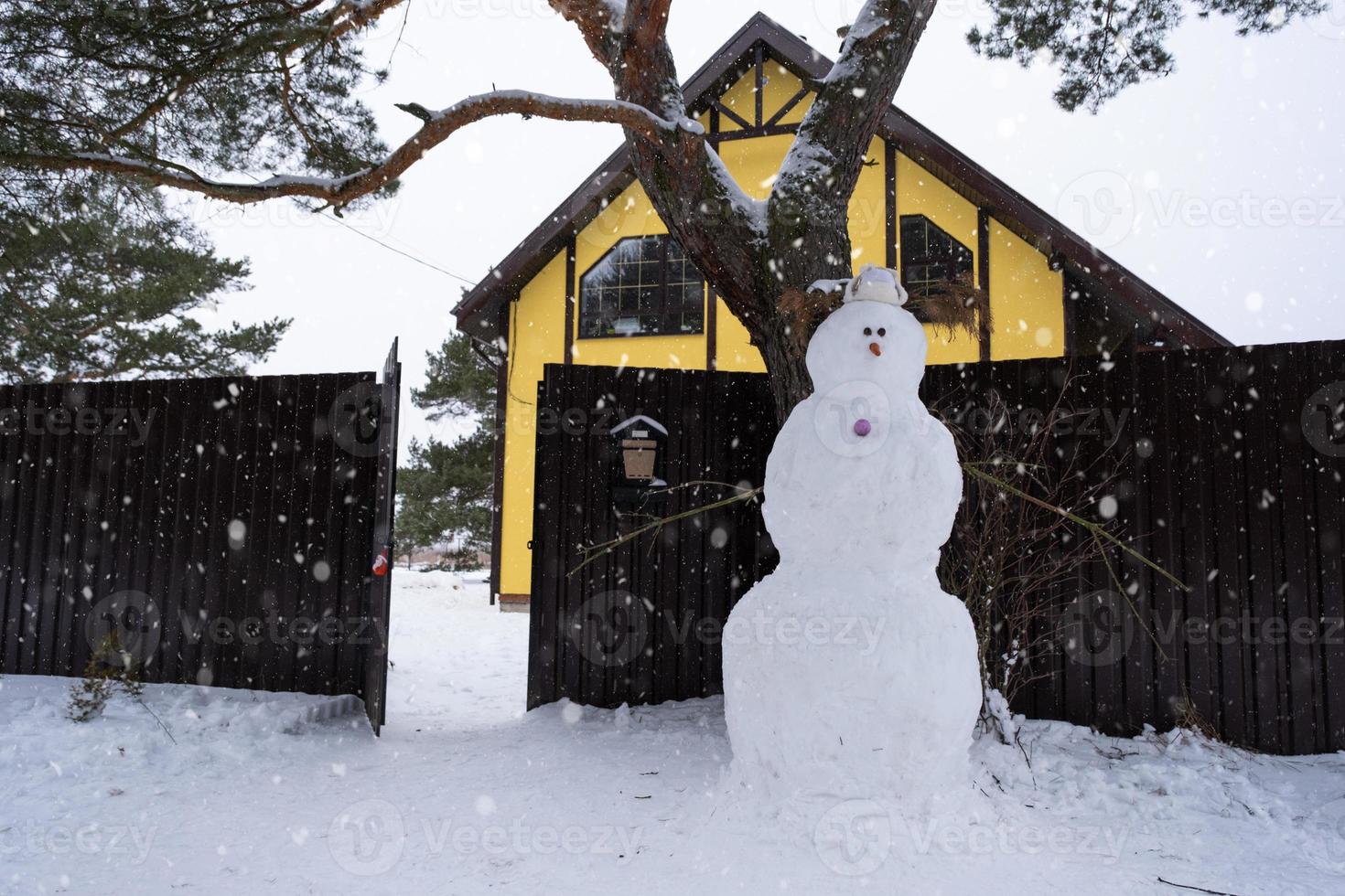 divertido muñeco de nieve con sombrero y bufanda en el fondo de una casa amarilla en el patio. invierno, entretenimiento de invierno, nevadas foto