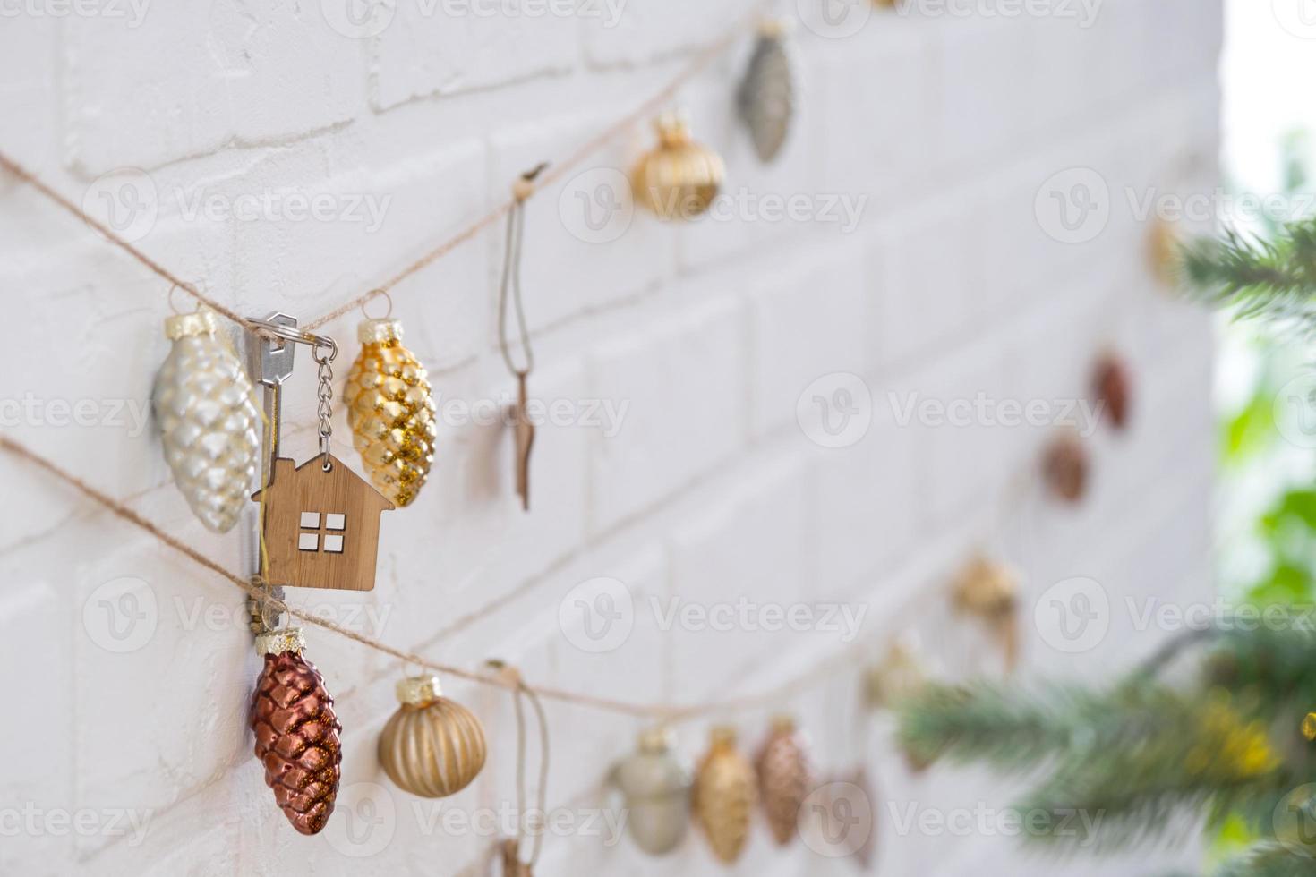 Key to house with keychain is hanging on garland of Christmas tree toys on wall. Gift for New Year, Christmas. Building, design, project, moving to new house, mortgage, rent and purchase real estate. photo