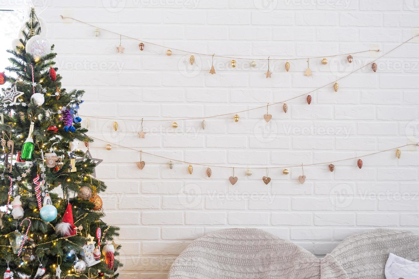 Christmas tree in white interior of a house with loft-style brick walls with garlands of glass toys on a rope. Glowing fairy lights decoration of the studio room. Cozy home in christmas and new year photo