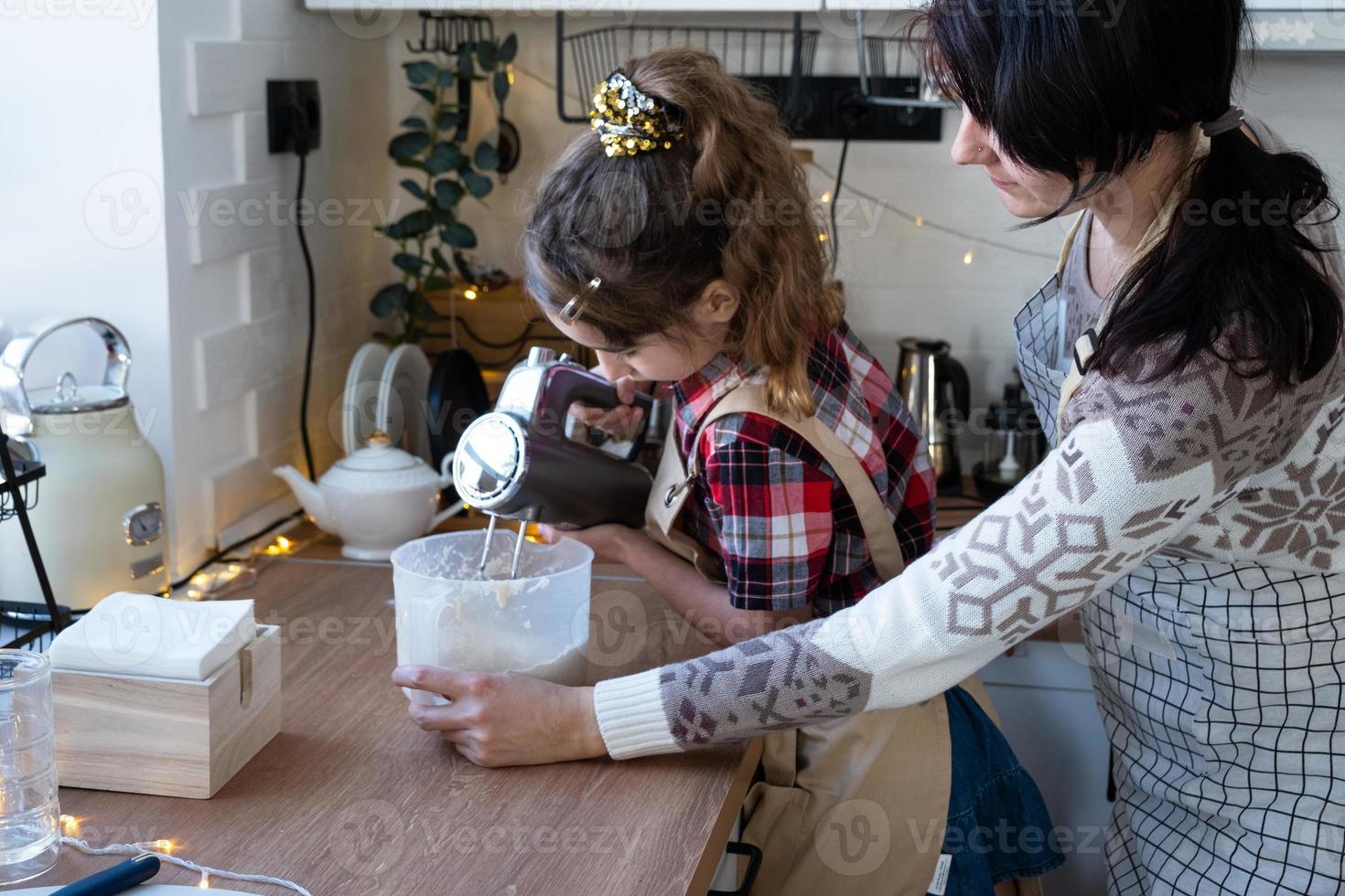Mom and daughter in the white kitchen knead the dough in the bowl for gingerbread and cookies with a blender for Christmas and new year. photo