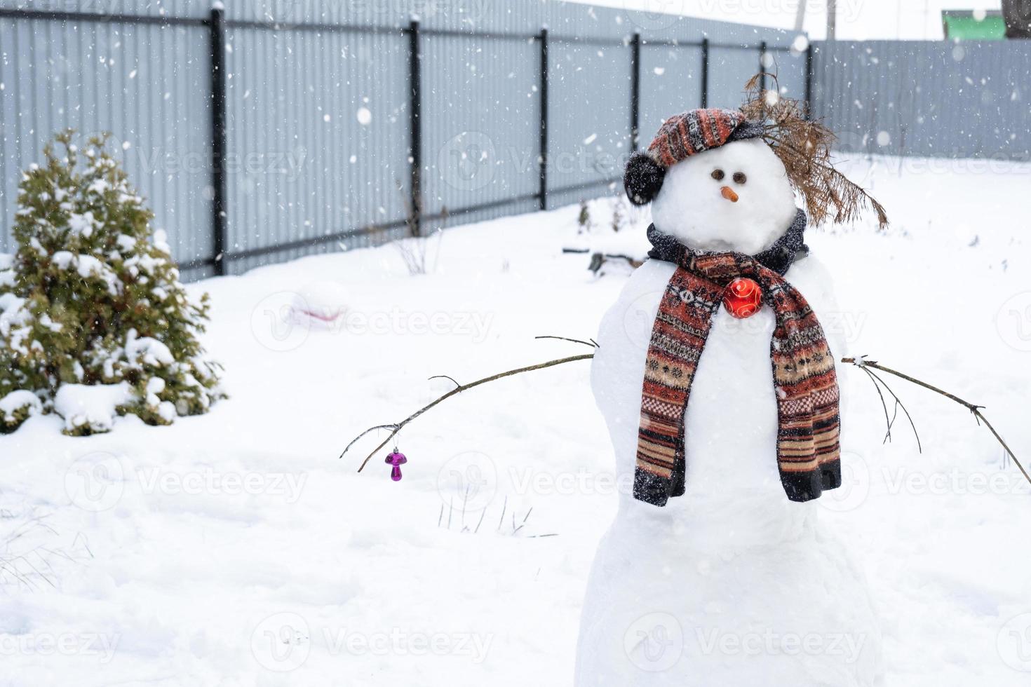 Funny snowman in a hat and scarf on the background of a yellow house in the yard. Winter, winter entertainment, snowfall photo