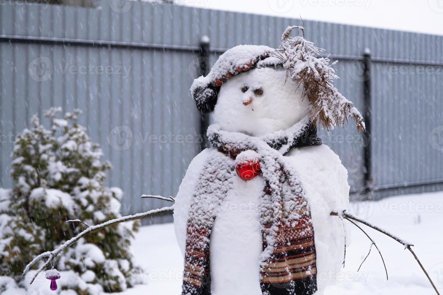 divertido muñeco de nieve con sombrero y bufanda en el fondo de una casa amarilla en el patio. invierno, entretenimiento de invierno, nevadas foto