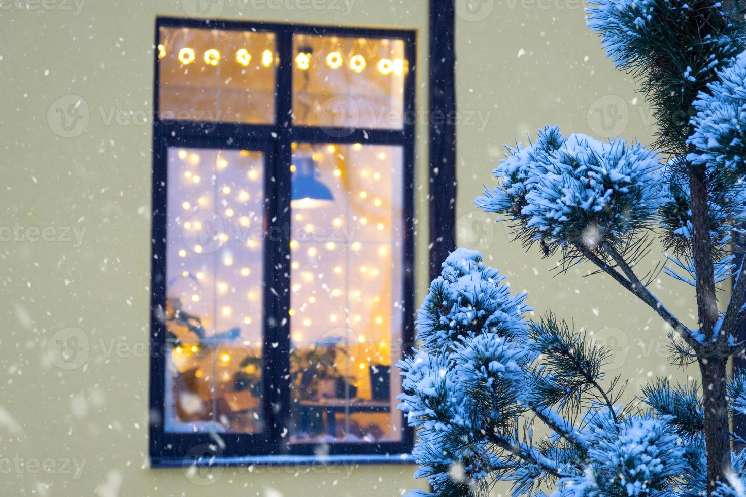 Cozy festive window of the house outside with the warm light of fairy lights garlands inside - celebrate Christmas and New Year in a warm home. Christmas tree, bokeh, snow on pine trees and snowfall photo