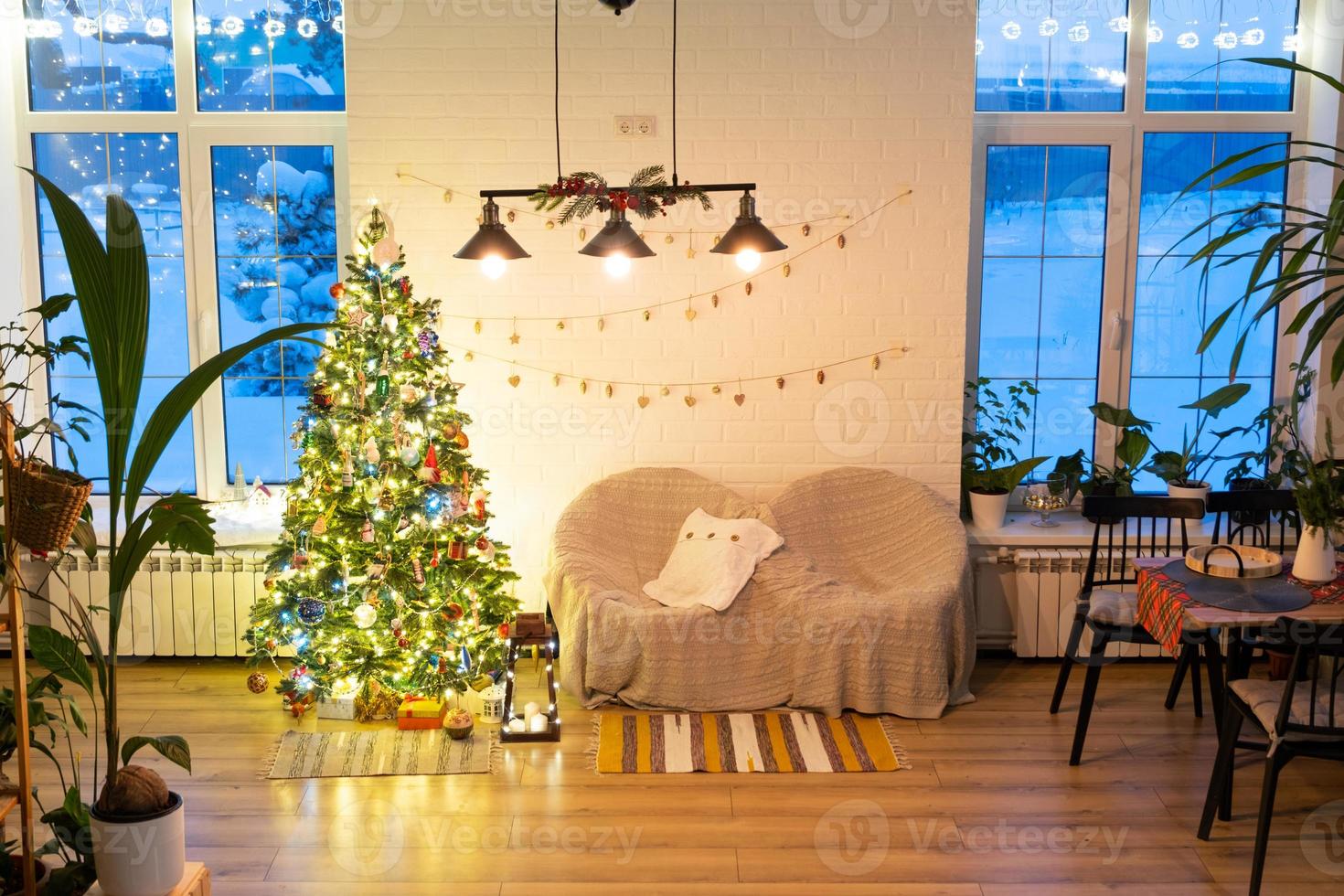 Christmas tree in white interior of a house with loft-style brick walls with garlands of glass toys on a rope. Glowing fairy lights decoration of the studio room. Cozy home in christmas and new year photo