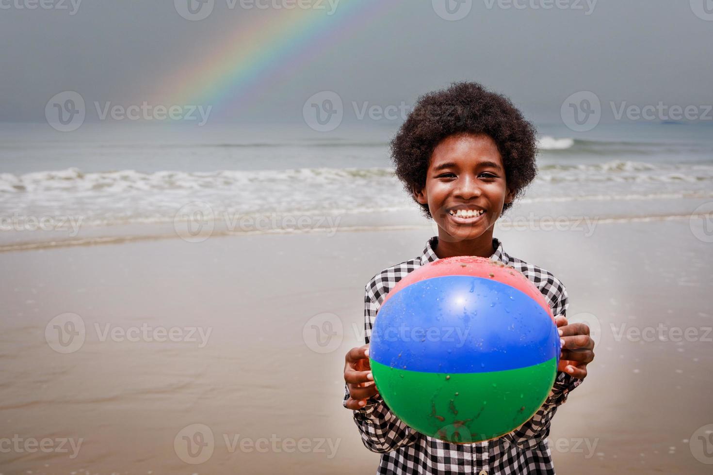 retrato de un niño afroamericano feliz sosteniendo una pelota de playa en una playa tropical. Diversidad étnica foto