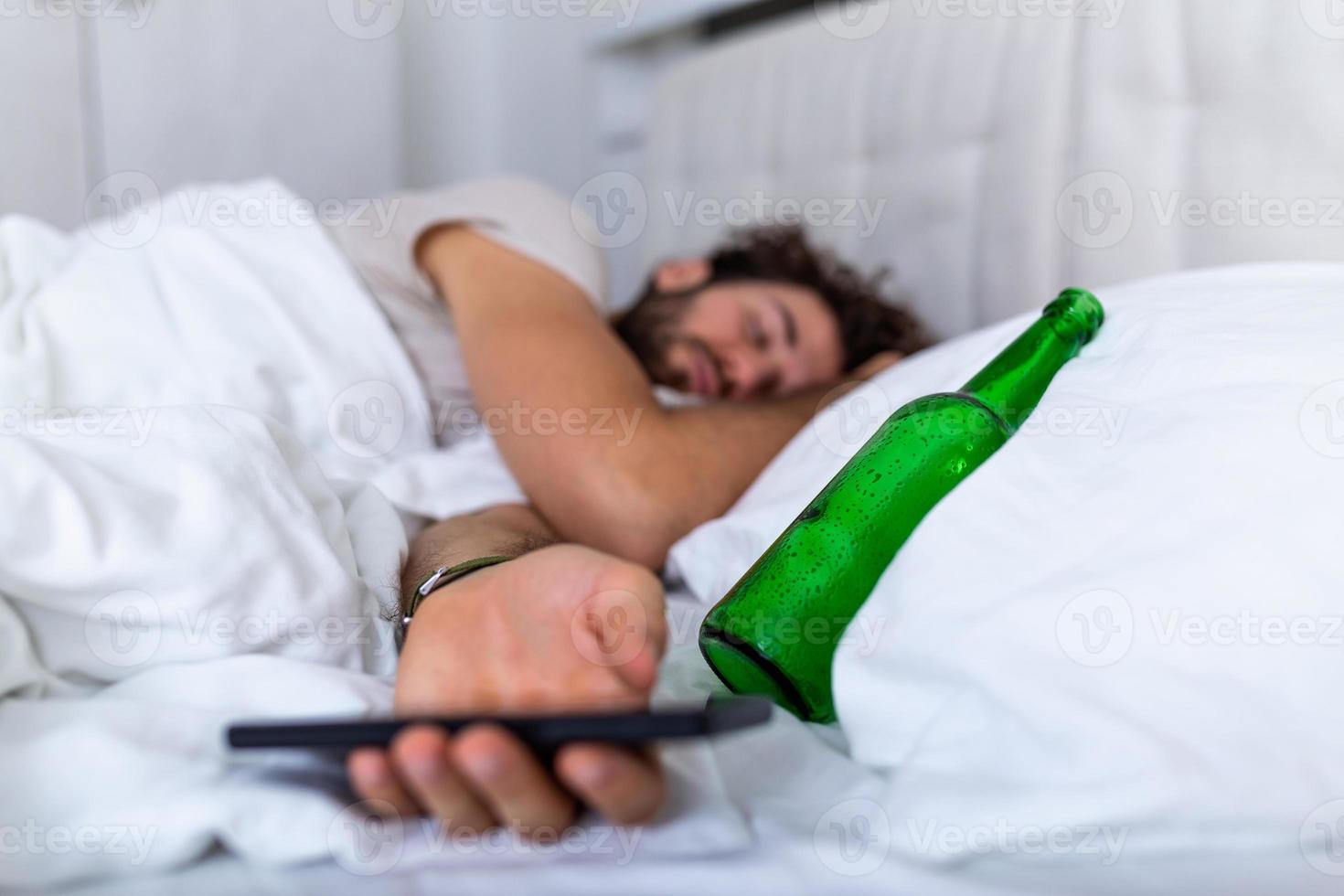 Texting while drunk concept. Young man lying in bed deadly drunken holding empty bottle of booze. intoxicated with alcohol . Alcoholism habitual drunkenness pernicious habit concept photo