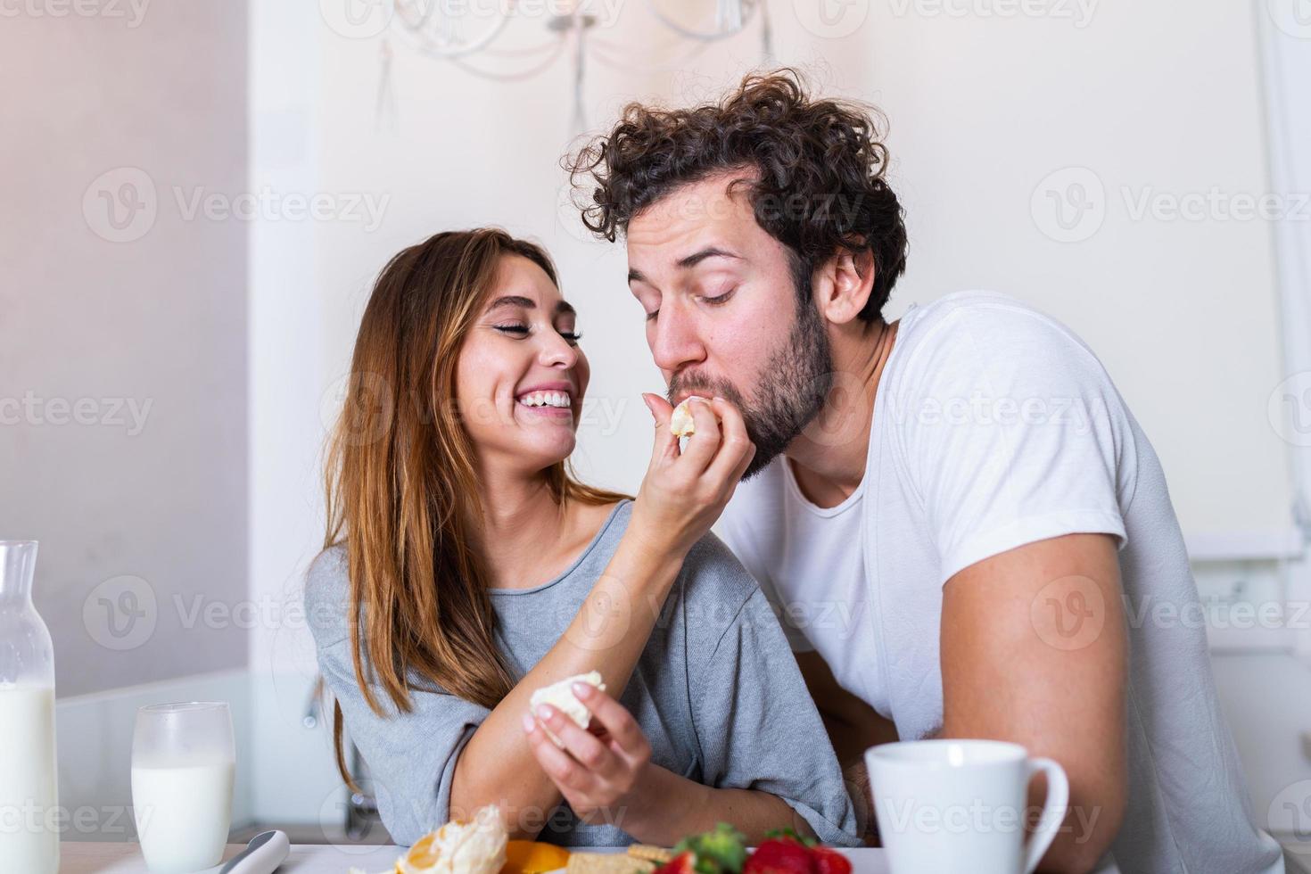 Beautiful young couple is looking at each other and feeding each other with smiles while cooking in kitchen at home. Loving joyful young couple embracing and cooking together photo