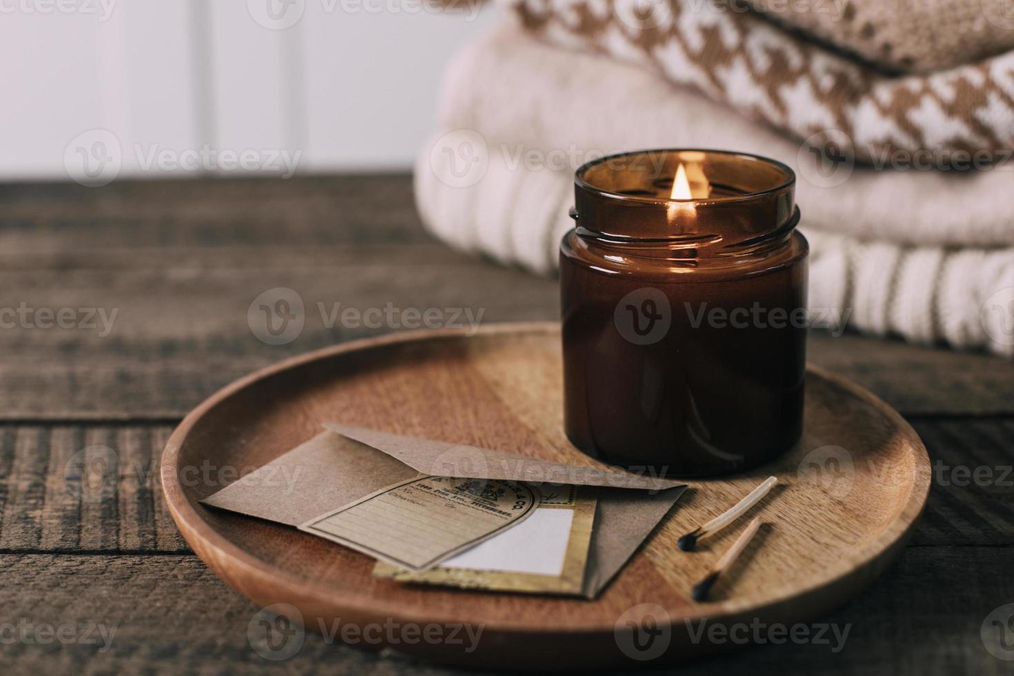 Burning candle in small amber glass jar with wooden wick, stack knitted season sweaters. Cozy lifestyle, hygge concept photo