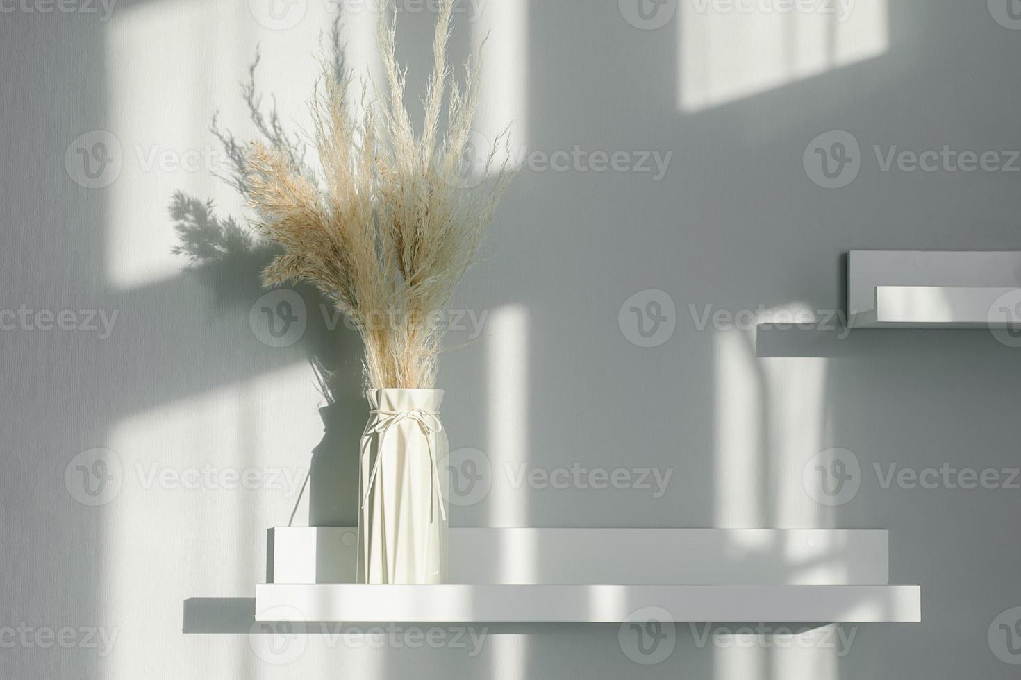 Home interior floral decor. Dried flowers in vase on white shelf. Background with shadow lights photo