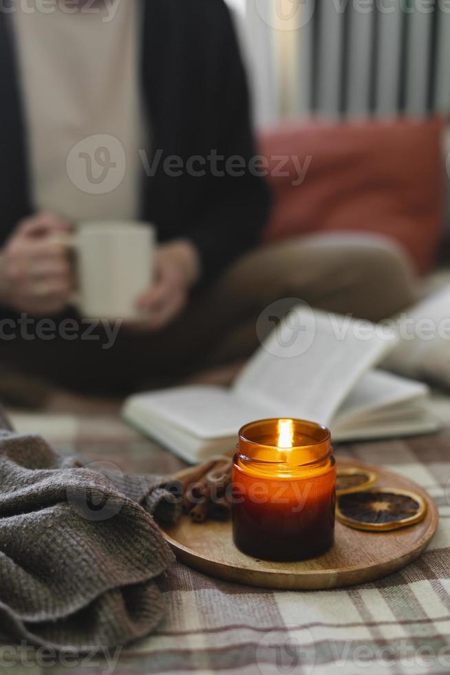 Burning candle on background of young woman holding mug coffee or tea, reading book while sitting in lotus pose on bed in cozy bedroom photo