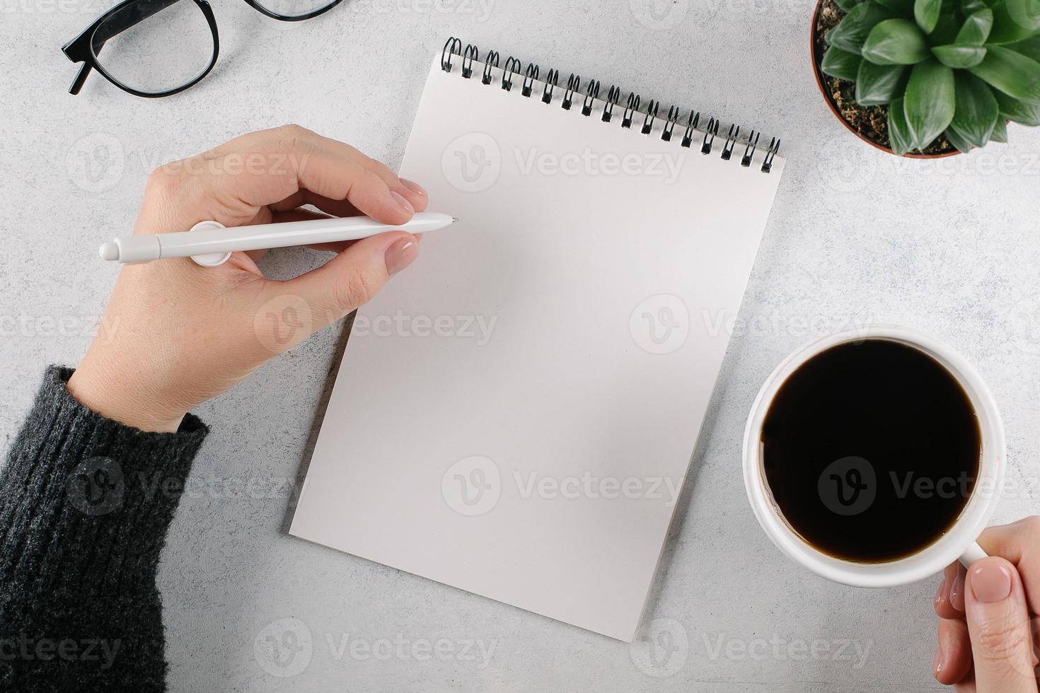 Womans hands writing to do list or plans in notebook and holding cup with coffee on office desk. Top view, copy space photo