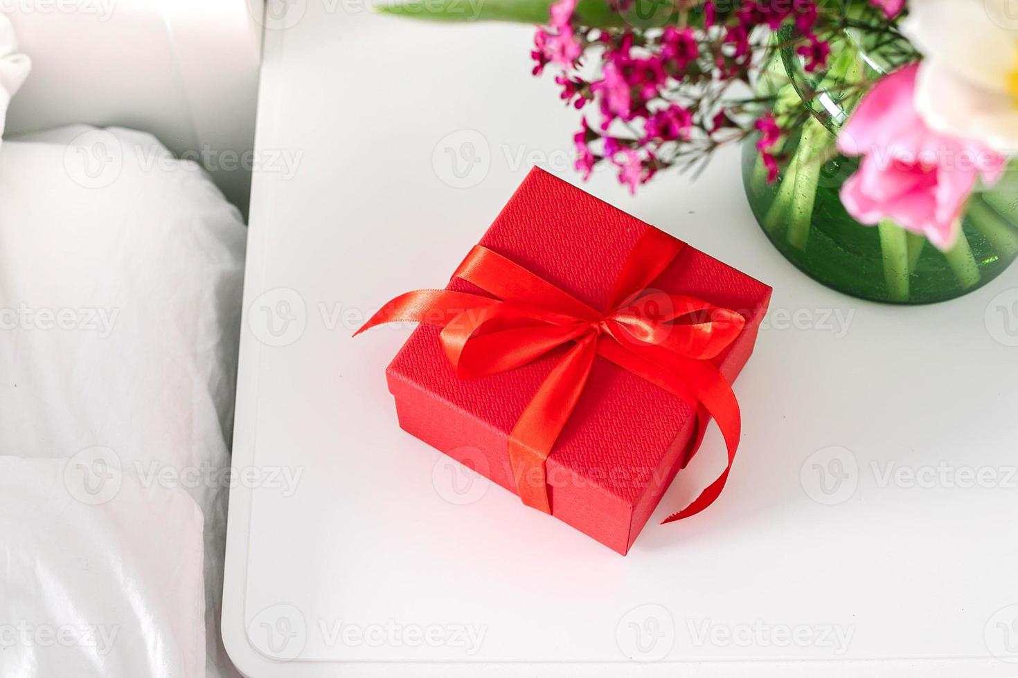 Bouquet of tulips and red gift box on the nightstand near the bed in the bedroom. Concept of womans day, mothers day photo