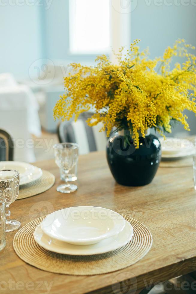 mesa de madera con platos y vasos vacíos. ajuste festivo de la mesa de pascua. ramas de mimosa en florero foto