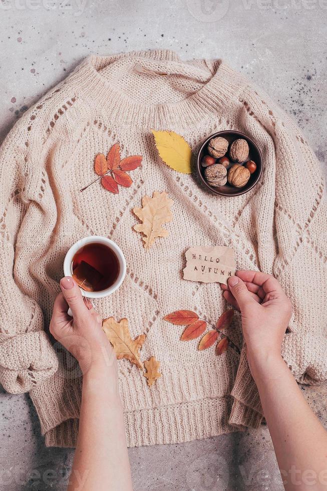 manos de mujer sosteniendo una taza de té y tarjeta de felicitación hola otoño. acogedor suéter beige. foto