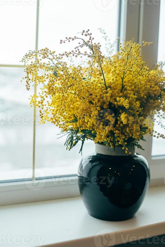 Bouquet of Mimosa flowers in blue vase on the windowsill next to the window. Happy women's day. photo