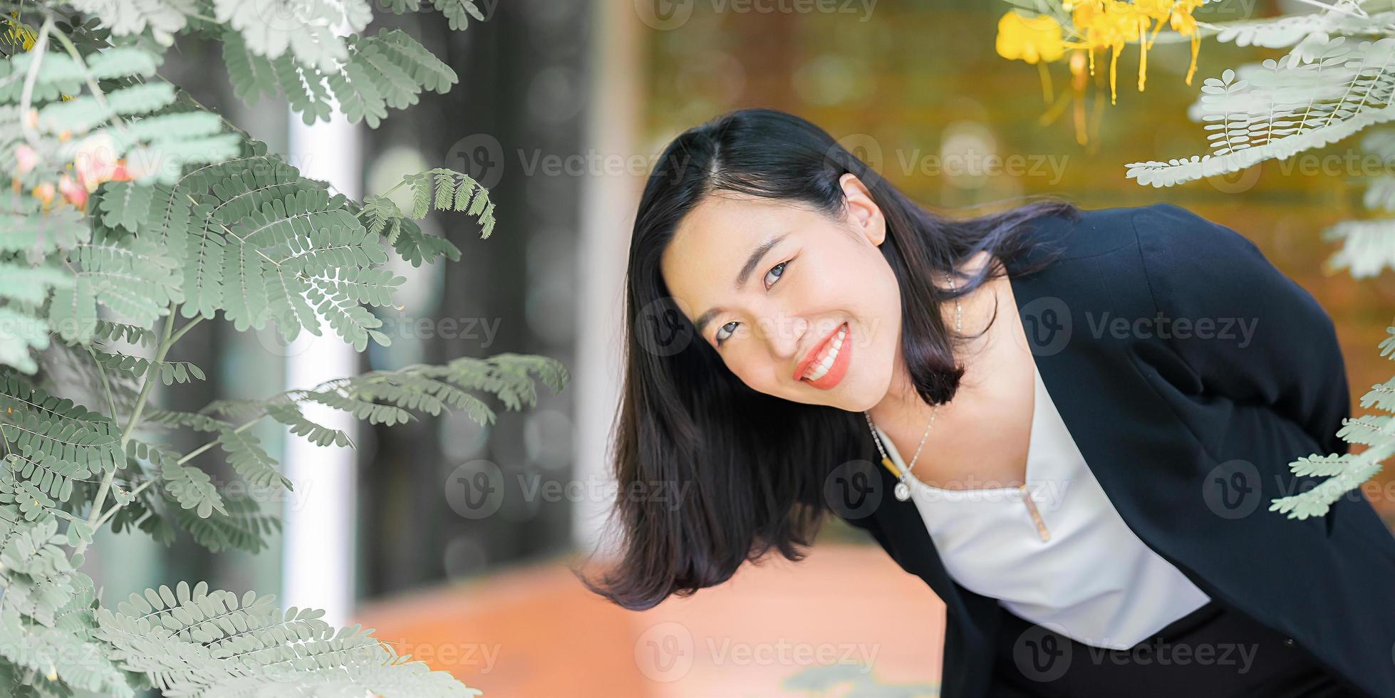 una joven asiática atractiva y confiada con traje negro de negocios está publicando, sonriendo frente a las hojas y el árbol con un estado de ánimo relajante en el jardín a la hora de la tarde. foto