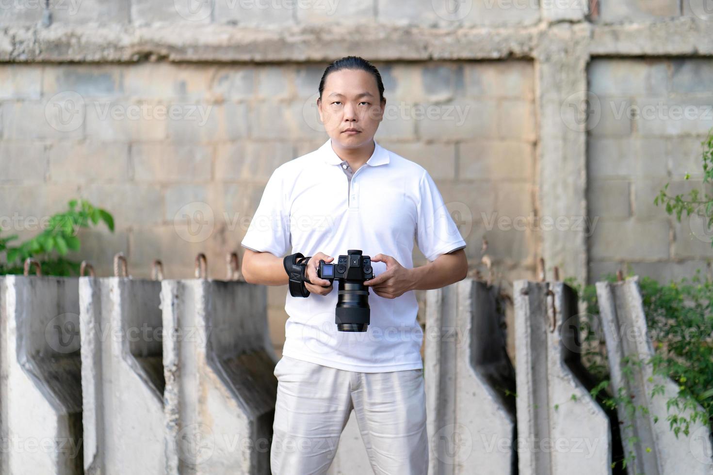 Asian man serious looks at the camera while holds th mirrorless camera medium format in the hand on the construction concret wall background. photo