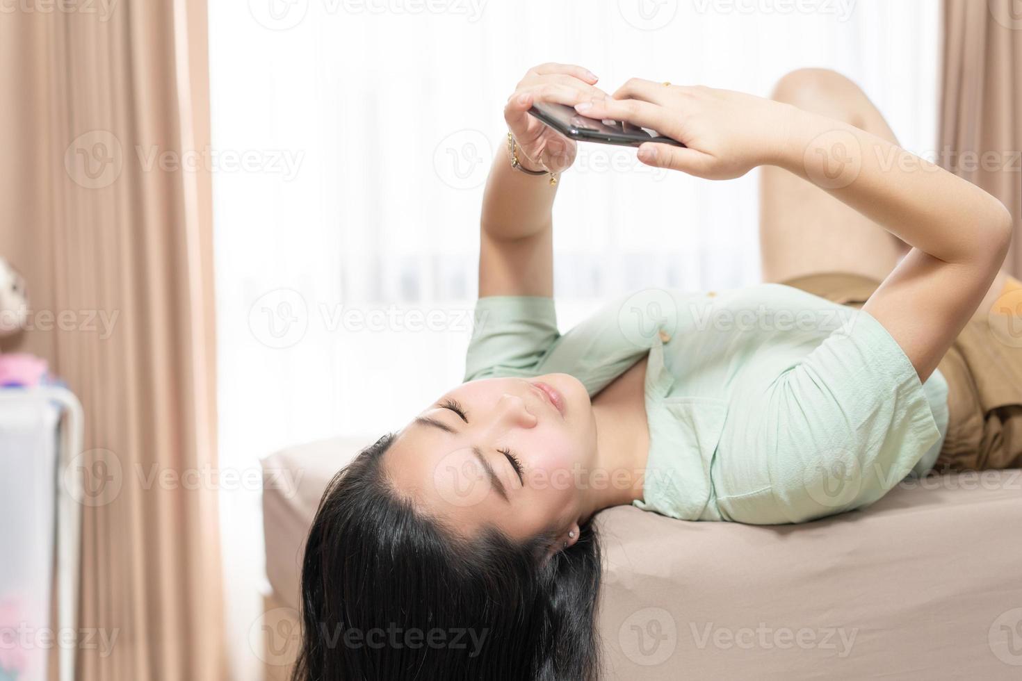 Beautiful Asian woman is lying down and playing mobile on the bed in her bedroom with a pastel green - brown color theme. photo