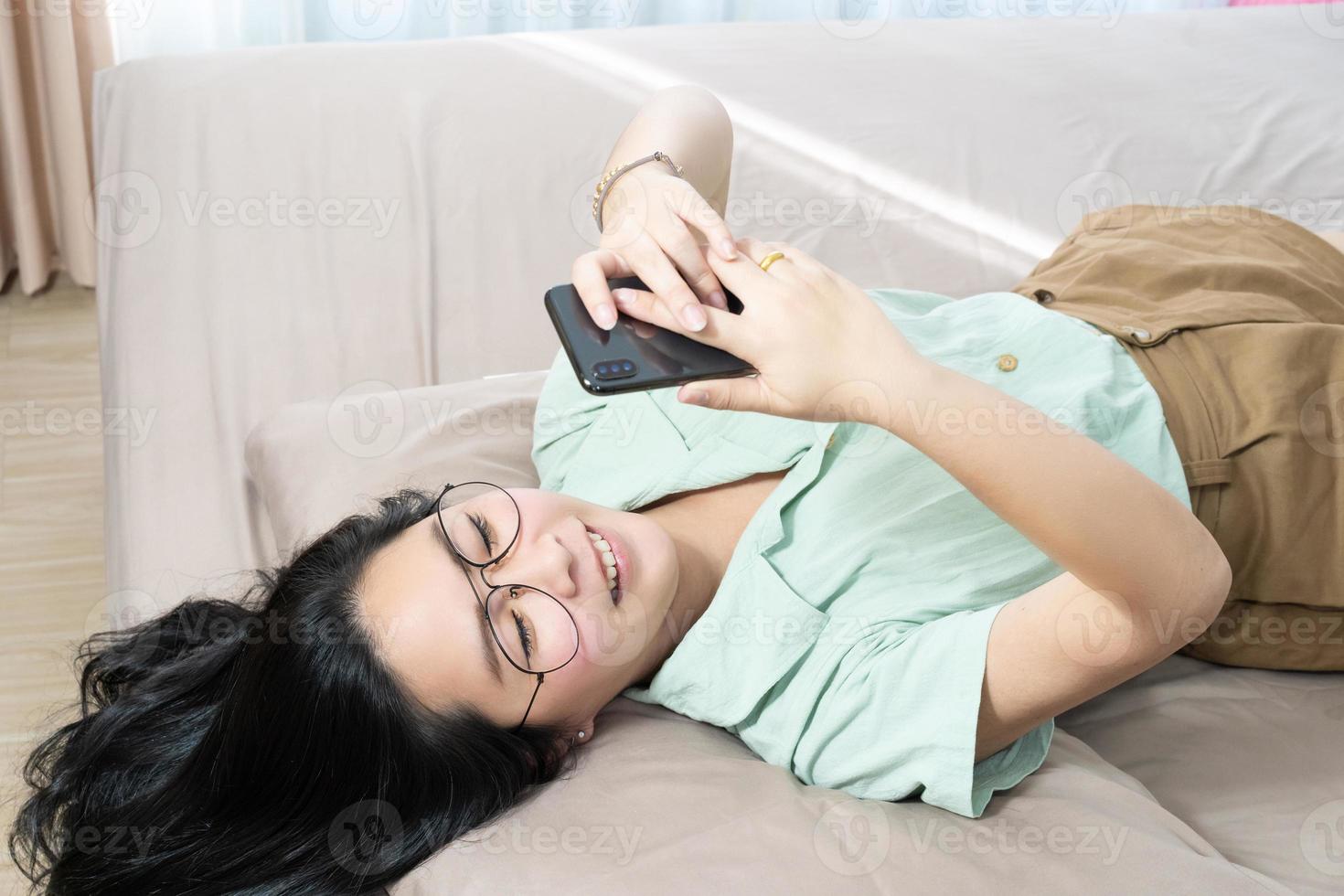 hermosas gafas mujer asiática yacen boca arriba, charlando, escribiendo y enviando mensajes de texto, móvil en su dormitorio con un tema de color verde pastel - marrón. foto