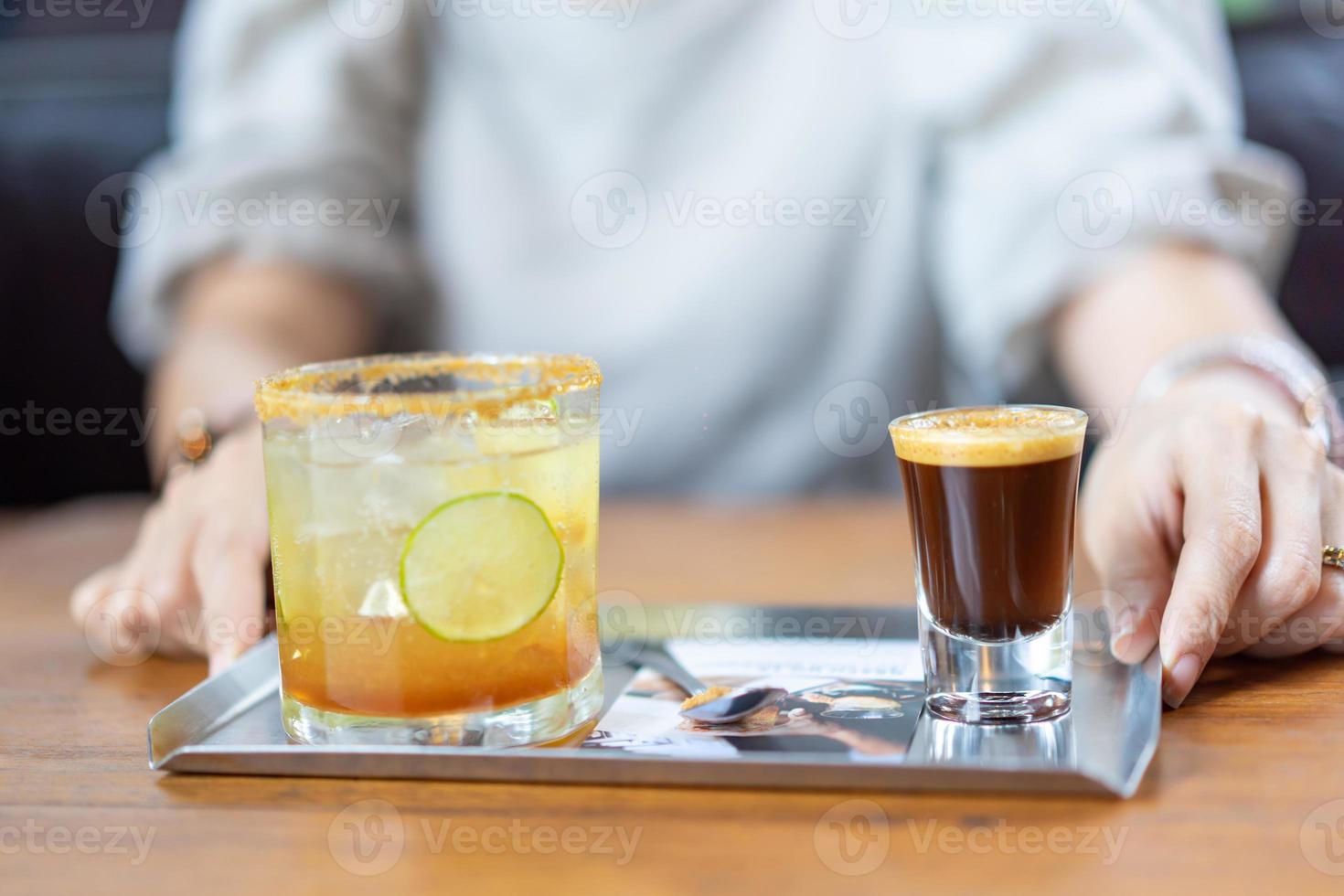 una combinación entre café recién hecho, limón, refresco de naranja en el plato de acero inoxidable en la mesa de madera frente a la mujer borrosa en el café. foto