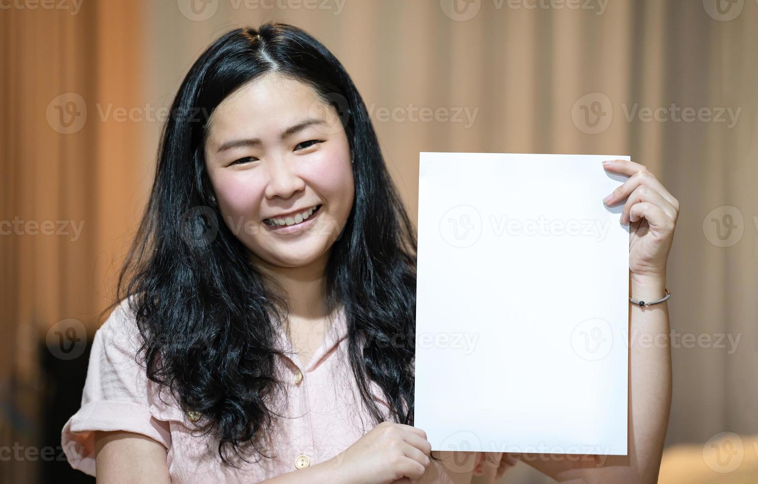 The Asian woman is holding and showing empty white A4 paper portrait - vertical angle on orange brown background. photo