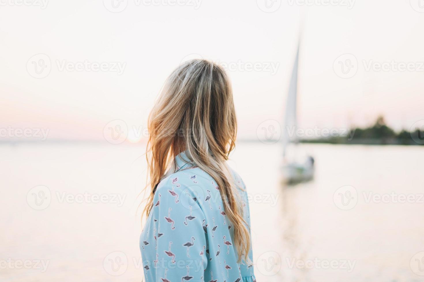 Beautiful blonde young woman in blue dress on pier on background of sailboat on the sunset photo