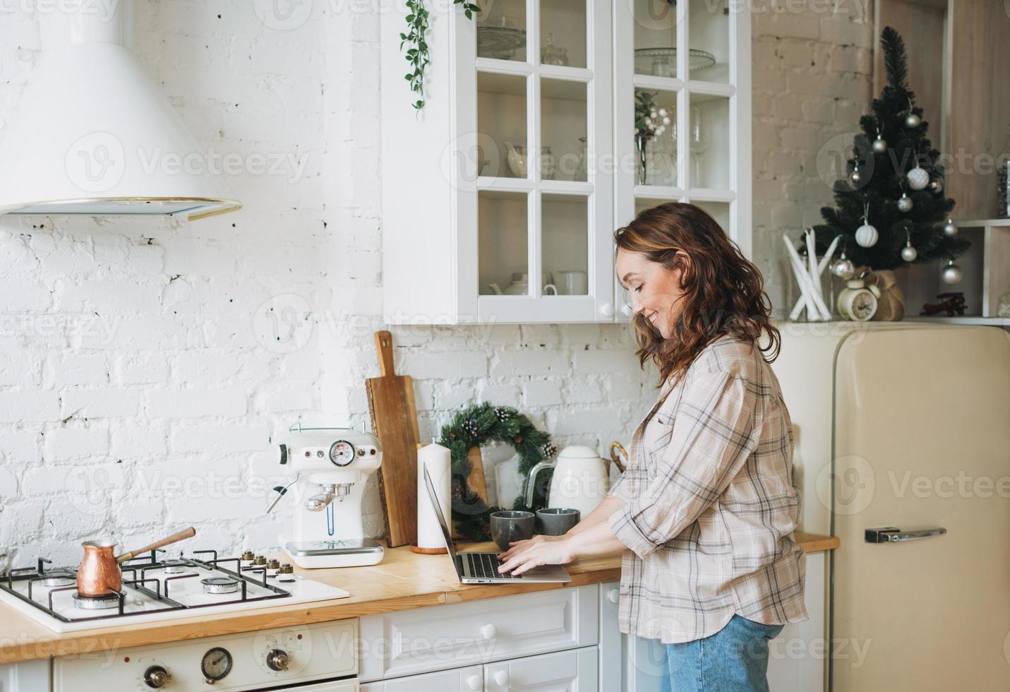 Attractive smiling woman with curly hair in plaid shirt with laptop near window at bright kitchen at home photo