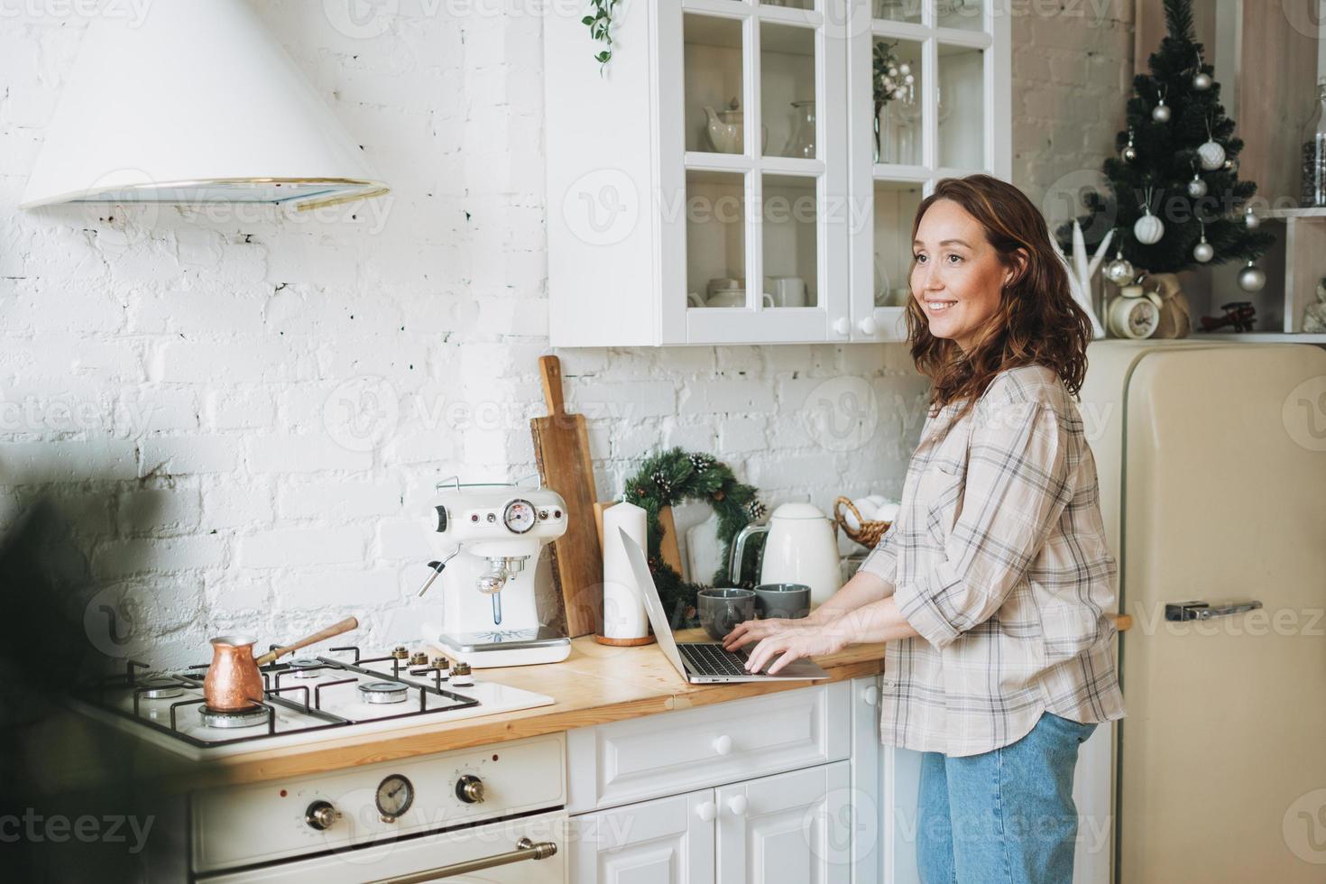 atractiva mujer sonriente con el pelo rizado en camisa a cuadros con portátil cerca de la ventana en la cocina luminosa en casa foto