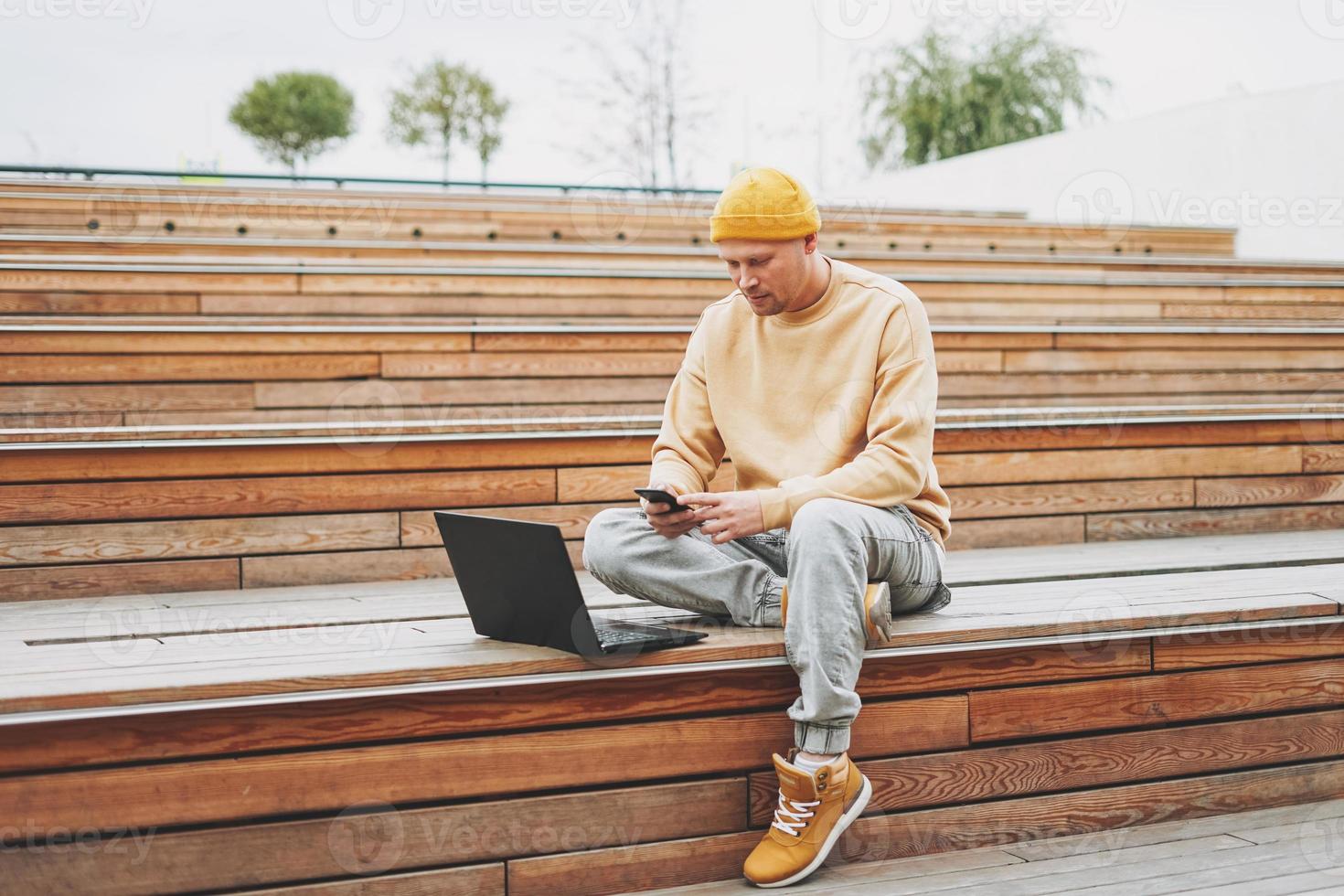 Stylish man hipster in yellow hat freelancer working on laptop in street city park photo