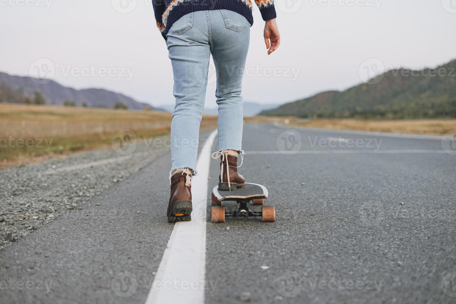 Crop photo of women's legs in jeans on longboard on road with the mountains background