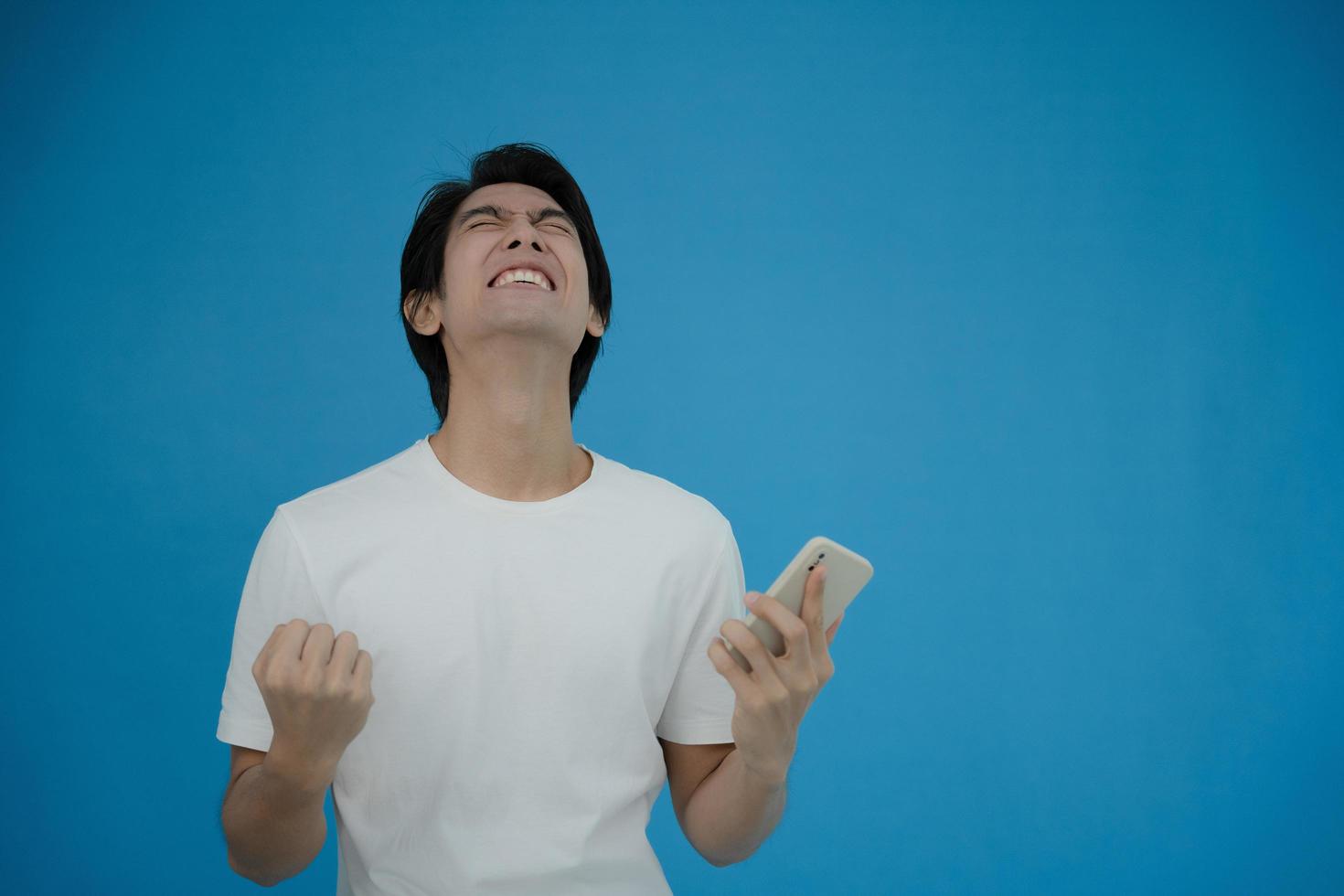 un hombre asiático genial emocionado y sorprendido por un teléfono inteligente con fondo azul. un hombre vio una venta en un sitio web de compras en línea o vio los resultados de la competencia de vítores del equipo de fútbol. foto