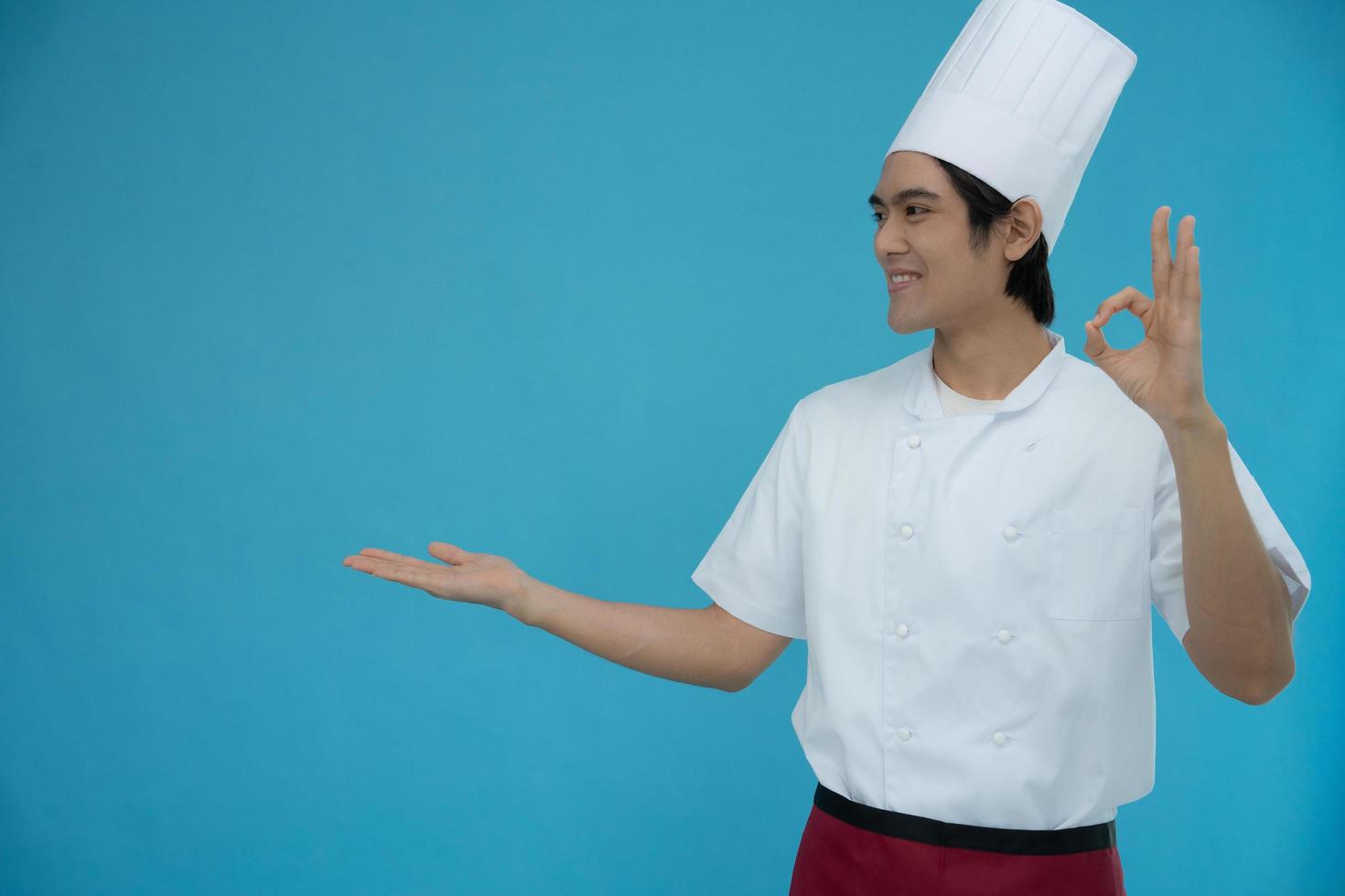 Portrait of a handsome Asian man wearing chef clothes on blue isolated background. Chef smiles and is happy with the food that is ok with the presentation. photo