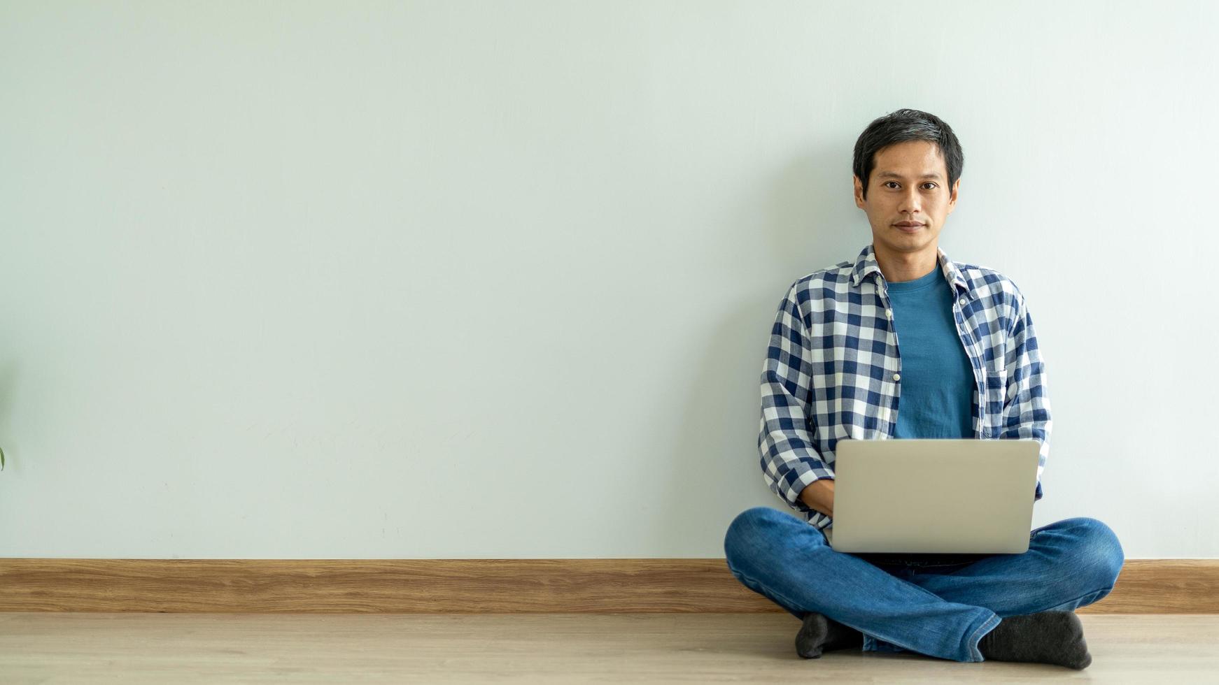 Asian man wearing a blue plaid shirt siting working on a laptop, relaxing on the floor of the room inside the house. Asian man ,Work from home concept photo