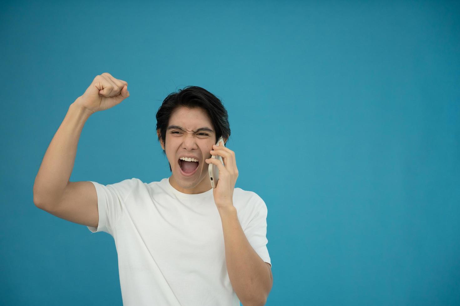 Cool asian man excited and surprised by smartphone on blue background. A man saw a sale on an online shopping website or See the results of the football team cheering competition. photo