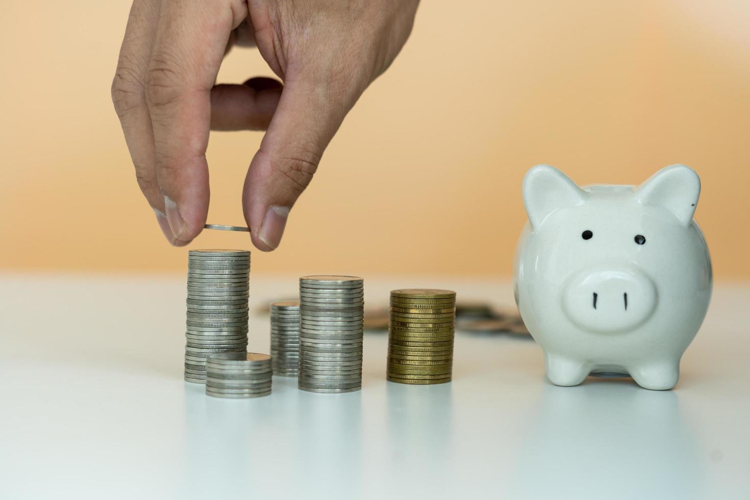 A man's hand is placing a silver coin stacked side by side with a white piggy bank. Man to collect savings as a business capital or savings money for the future. Concept saving money photo