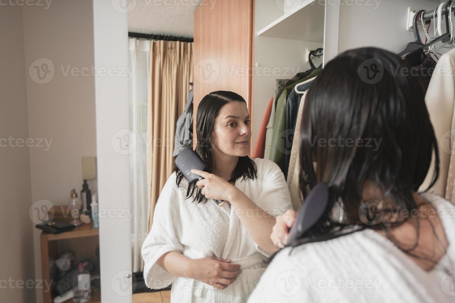 woman in a white bathrobe combing her hair with a brush in front of the mirror on her closet door in her bedroom photo