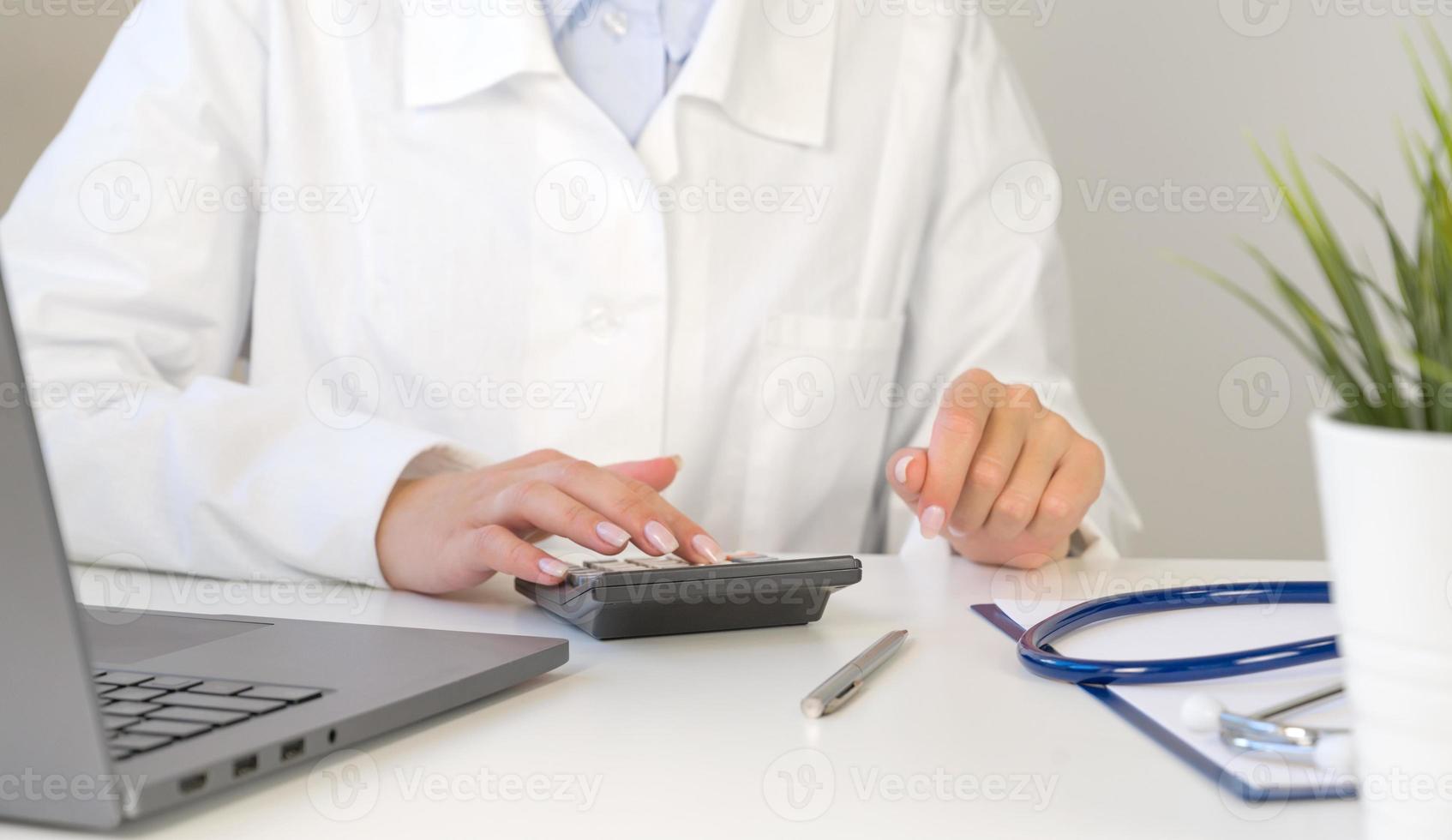 Front view at unrecognizable doctor using calculator with laptop and stethoscope near. Medical insurance concept photo