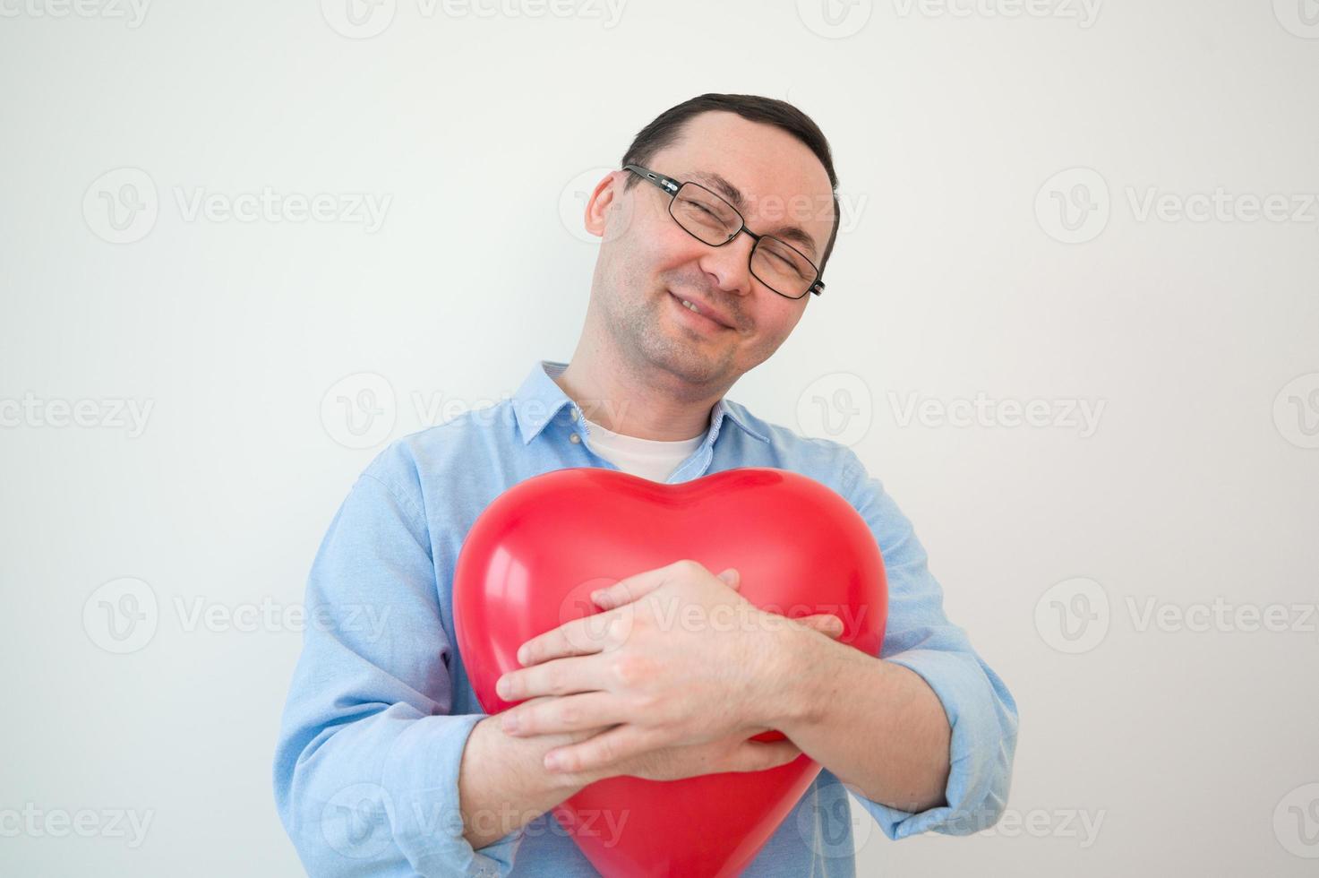 Close up photo of a charming man, who is laughing and holding a red heart-shaped balloon in his hands against white wall